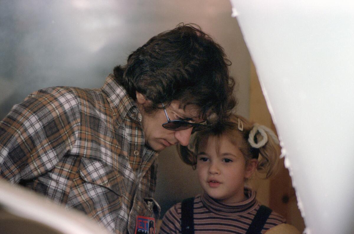 Steven Spielberg wears aviator sunglasses as he leans down to a young Drew Barrymore has her hair in pigtails.