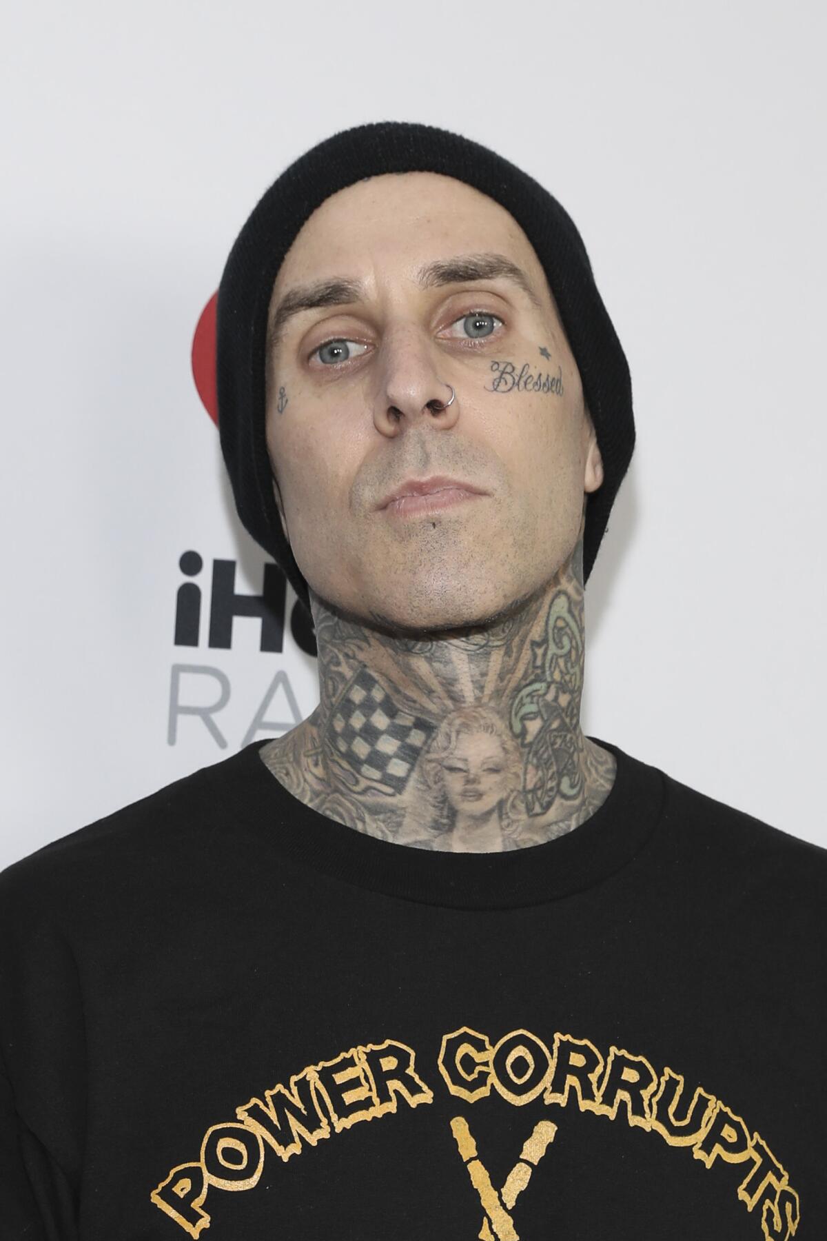 A man, wearing a black T-shirt and beanie, poses for photos. He has tattoos on his face and neck. 