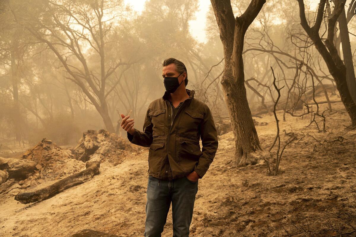 Gavin Newsom, wearing a mask, stands on a hillside in the haze of wildfire smoke.