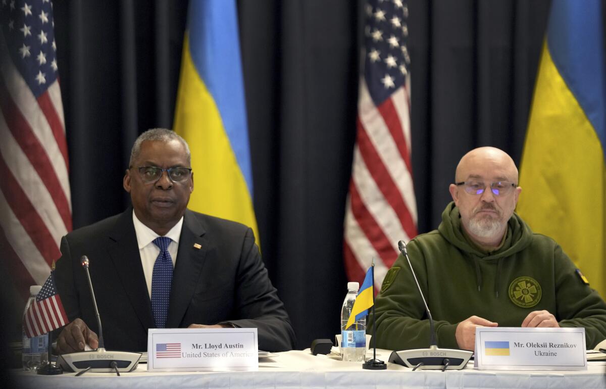 Two men sit side by side with flags behind them