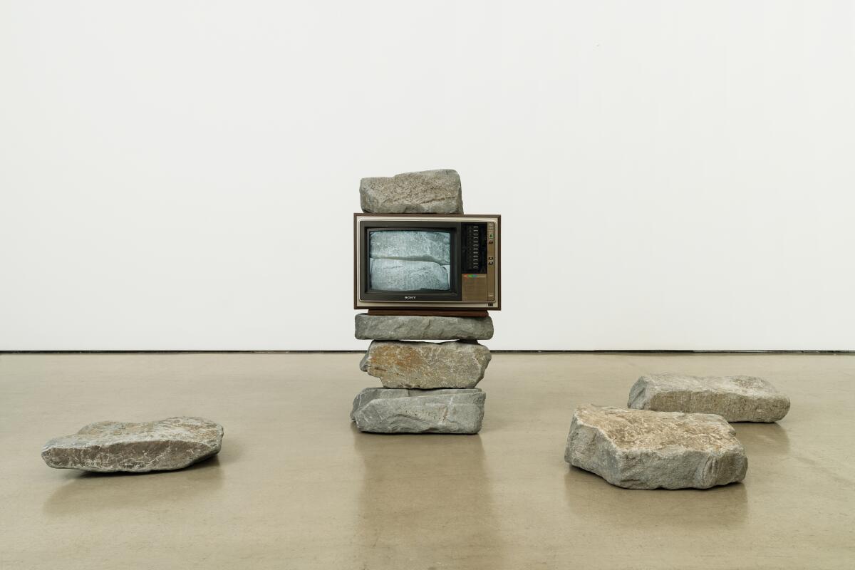 A television monitor sits on top of a stack of hefty stones, surrounded by additional scattered rocks.