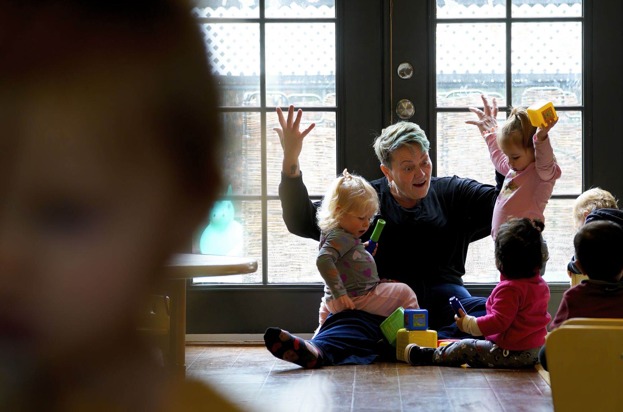 A woman sits on the floor in front of glass doors, gesturing animatedly to four toddlers.