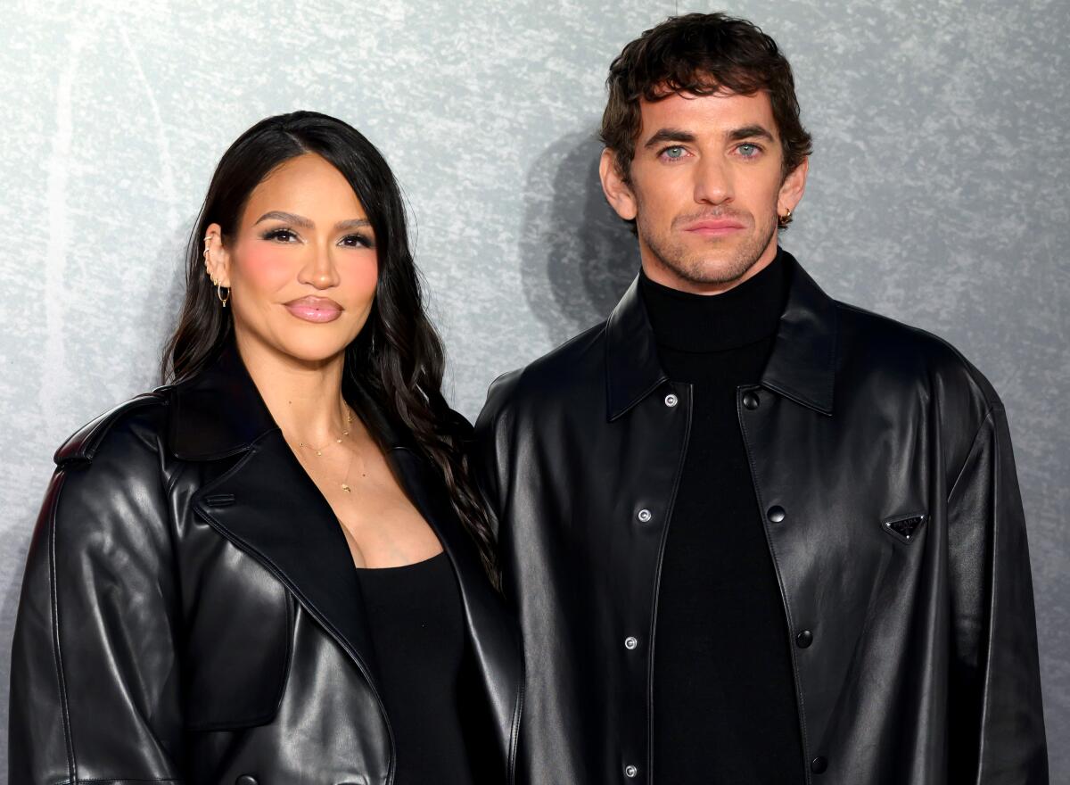 Cassie and Alex Fine in matching black leather coats pose in front of a gray patterned backdrop