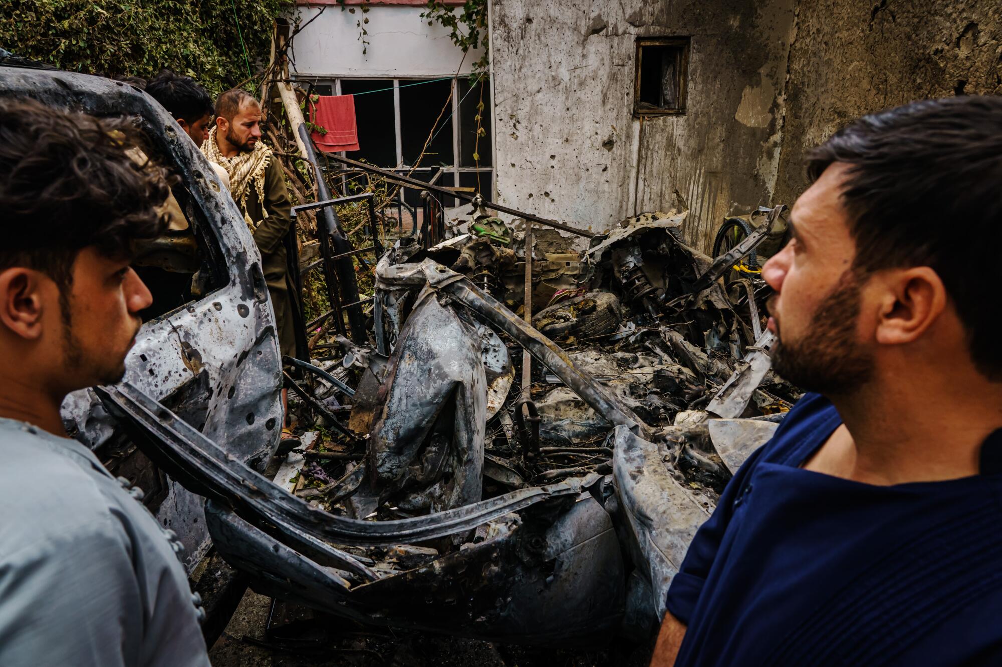 Relatives and neighbors of the Ahmadi family gather around the incinerated husk of a vehicle.