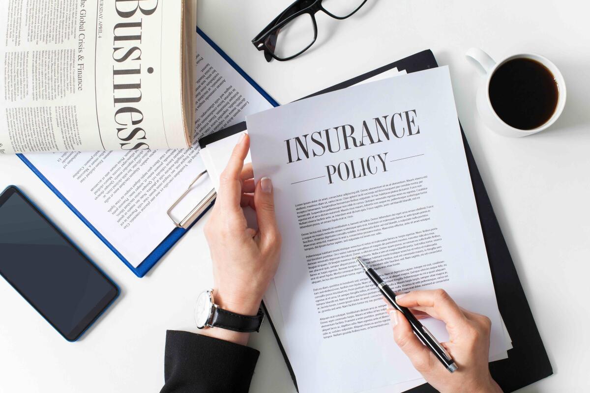 The hands of a businesswoman examining an insurance policy with a business newspaper, eyeglasses, and coffee on a white desk.