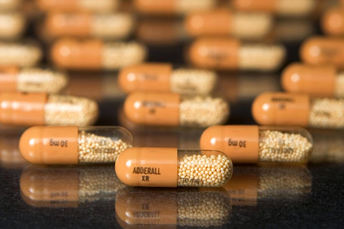 More than a dozen orange capsules labeled "Adderall XR" and "30 mg" arranged on a dark reflective surface. 