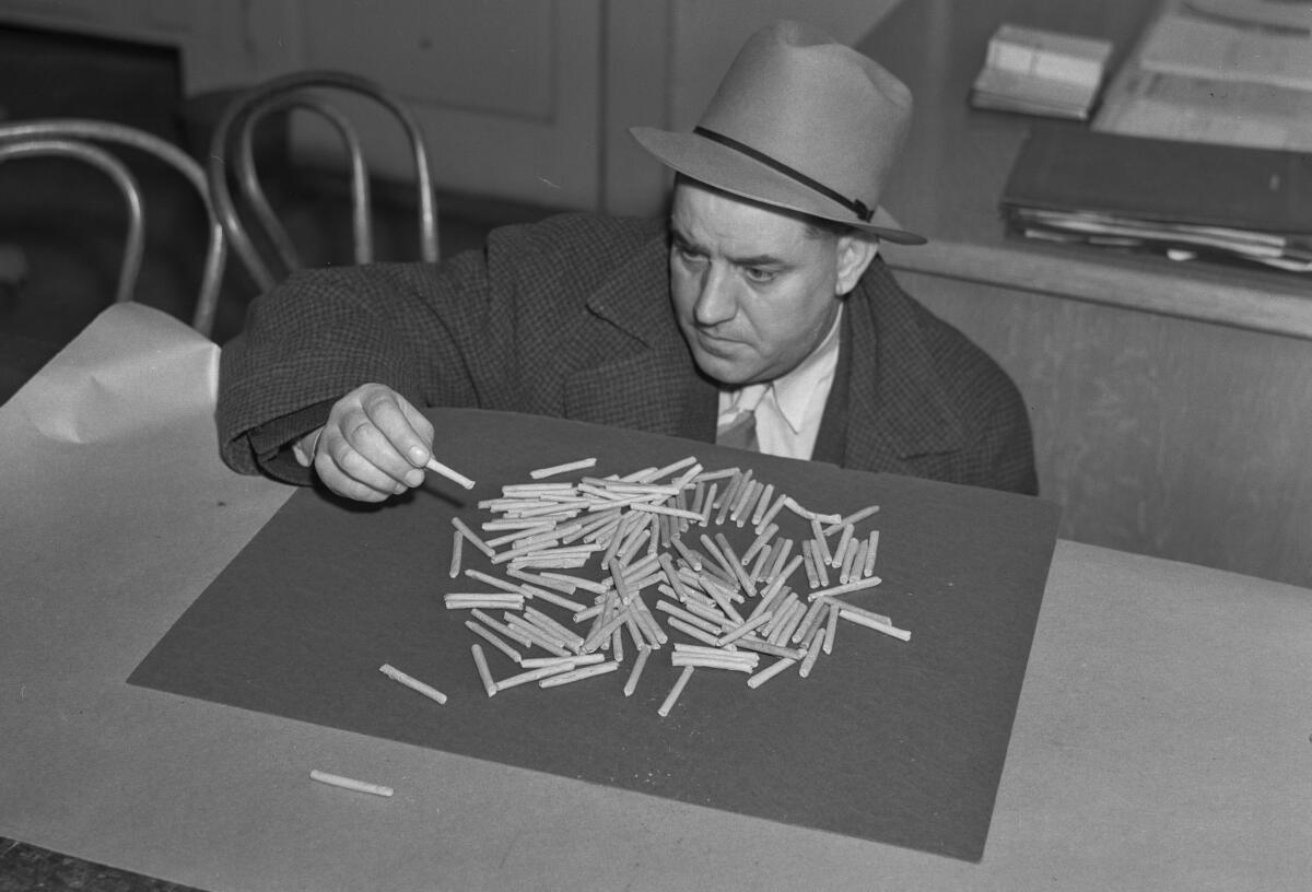 A man in a hat, suit, tie and overcoat leans over a pile of joints on a desktop.