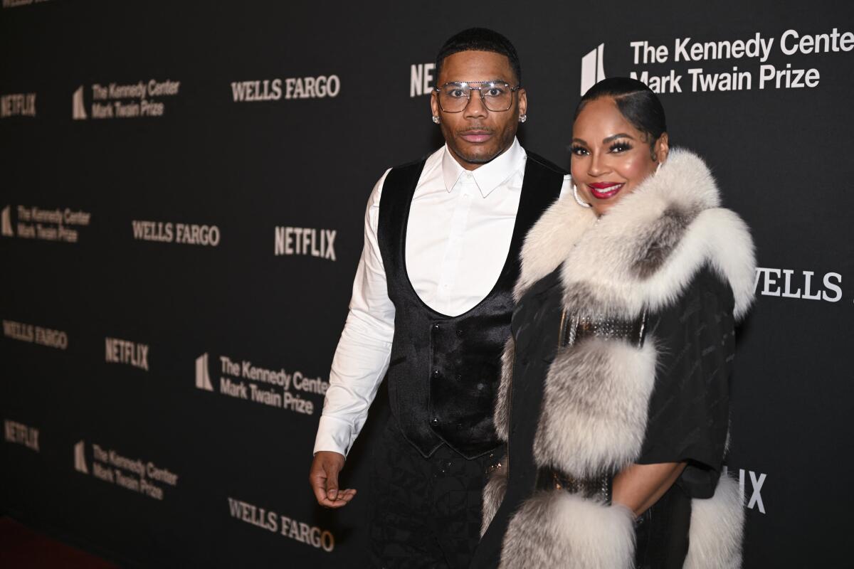 Nelly wearing black pants and a black scoop-neck vest over a white button down and Ashanti wearing a fur over black