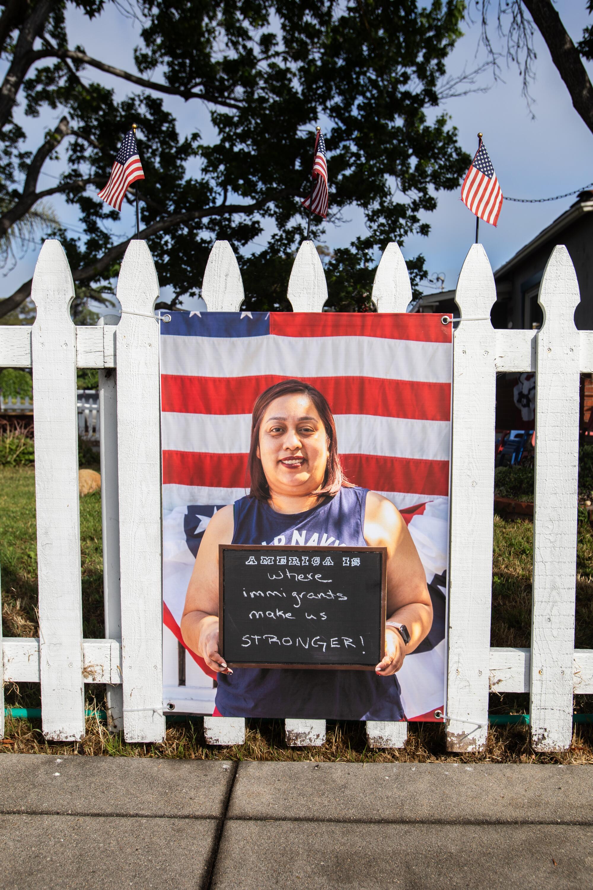  A portrait of a woman holding a sign in front of an American flag hangs on a white picket fence 