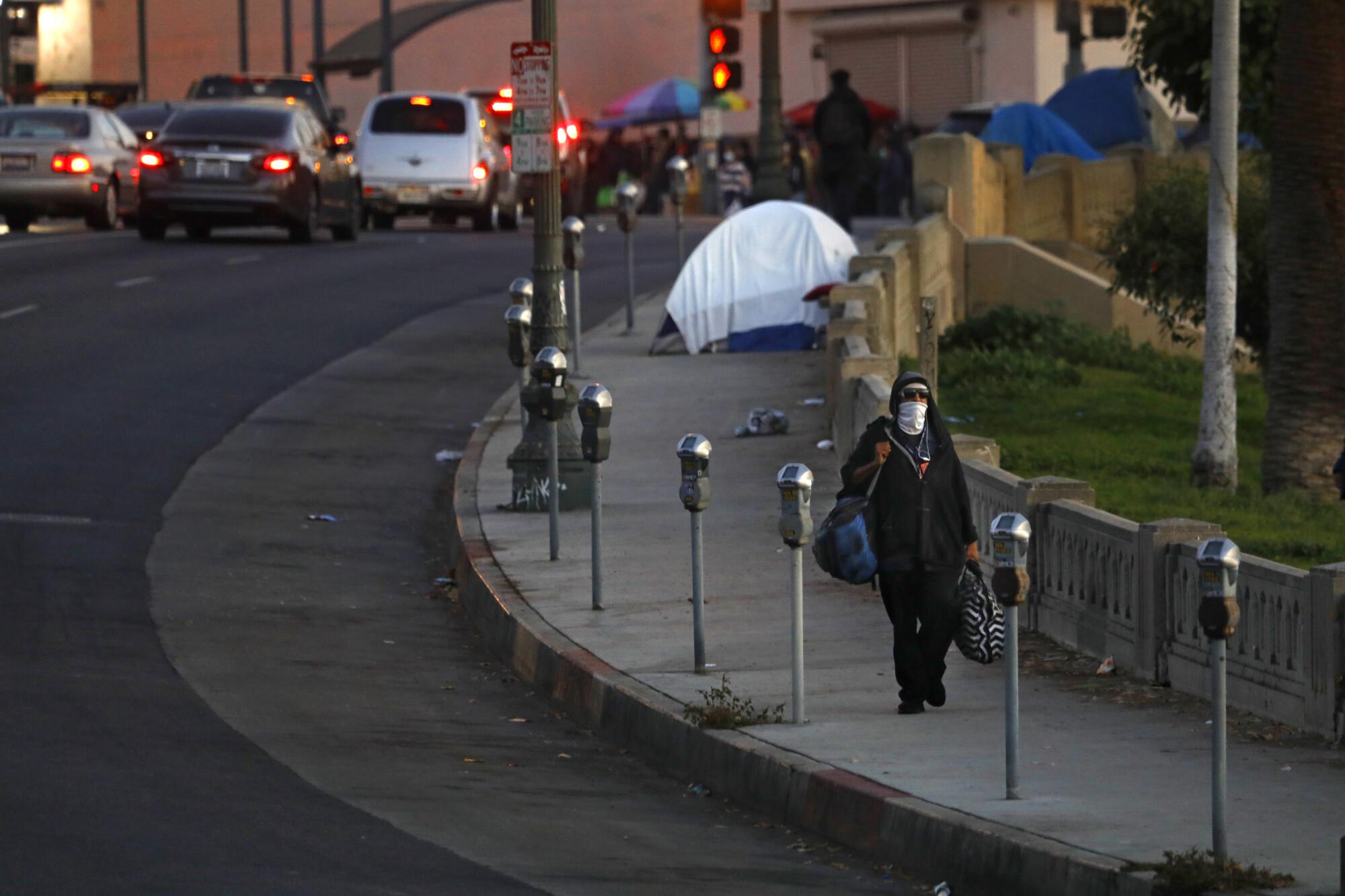 A person in a face covering walks on a city sidewalk with a tent and vendors' carts in the background
