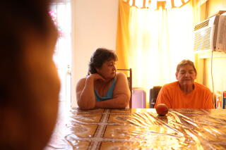 A woman rests her head on a hand, looking at a woman seated beside her at a table, as sun seeps through a curtain behind them