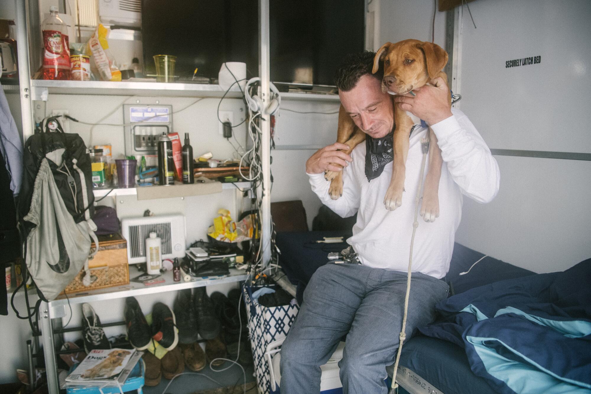 A puppy is draped around a man's neck and they are surrounded by his belongings. 