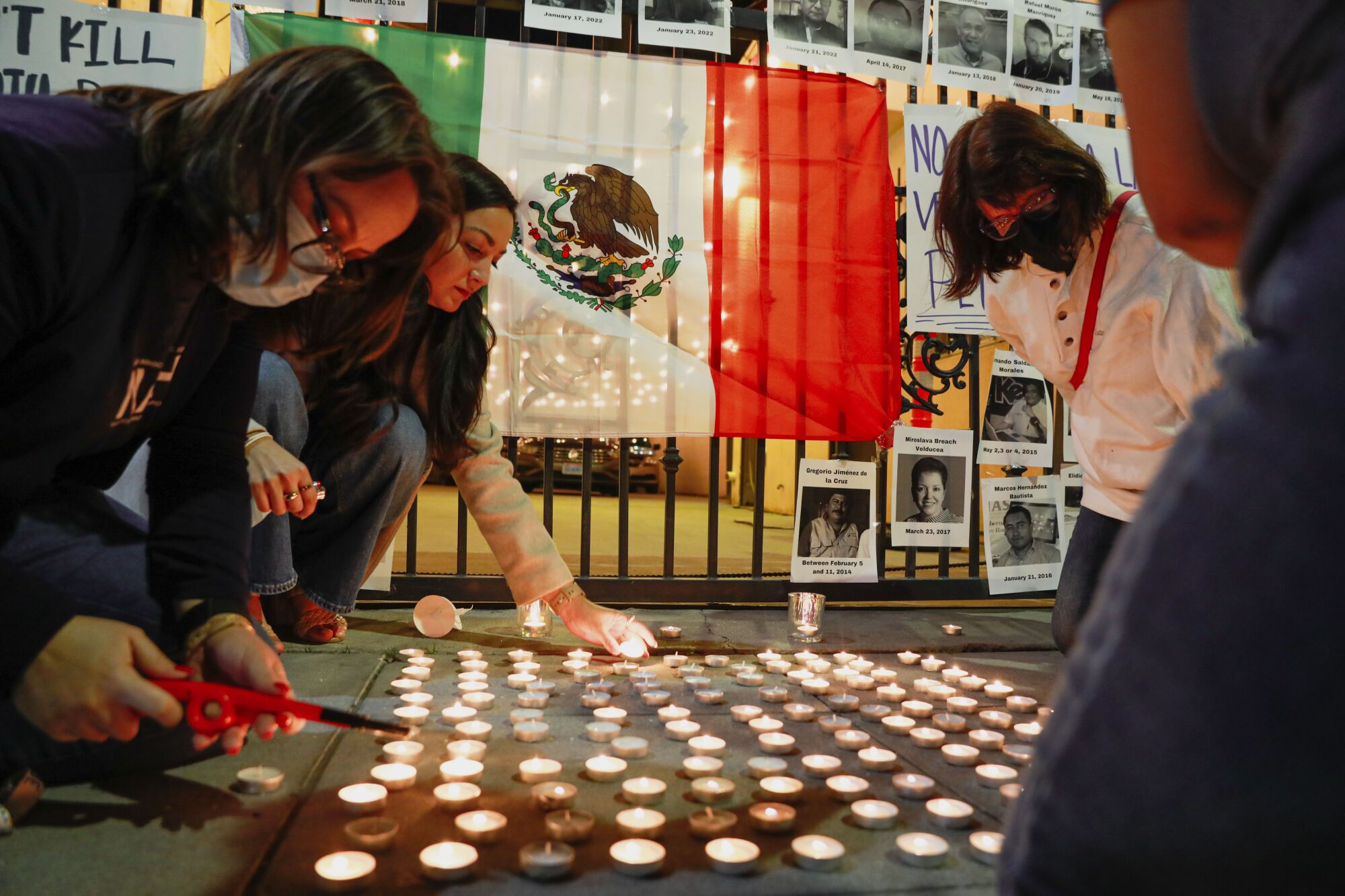 People light candles on a sidewalk next to a Mexican flag.