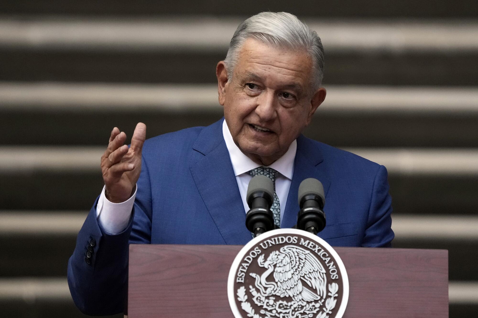 A man with gray hair, in a blue suit, gestures with one hand while speaking at a lectern with a government seal 