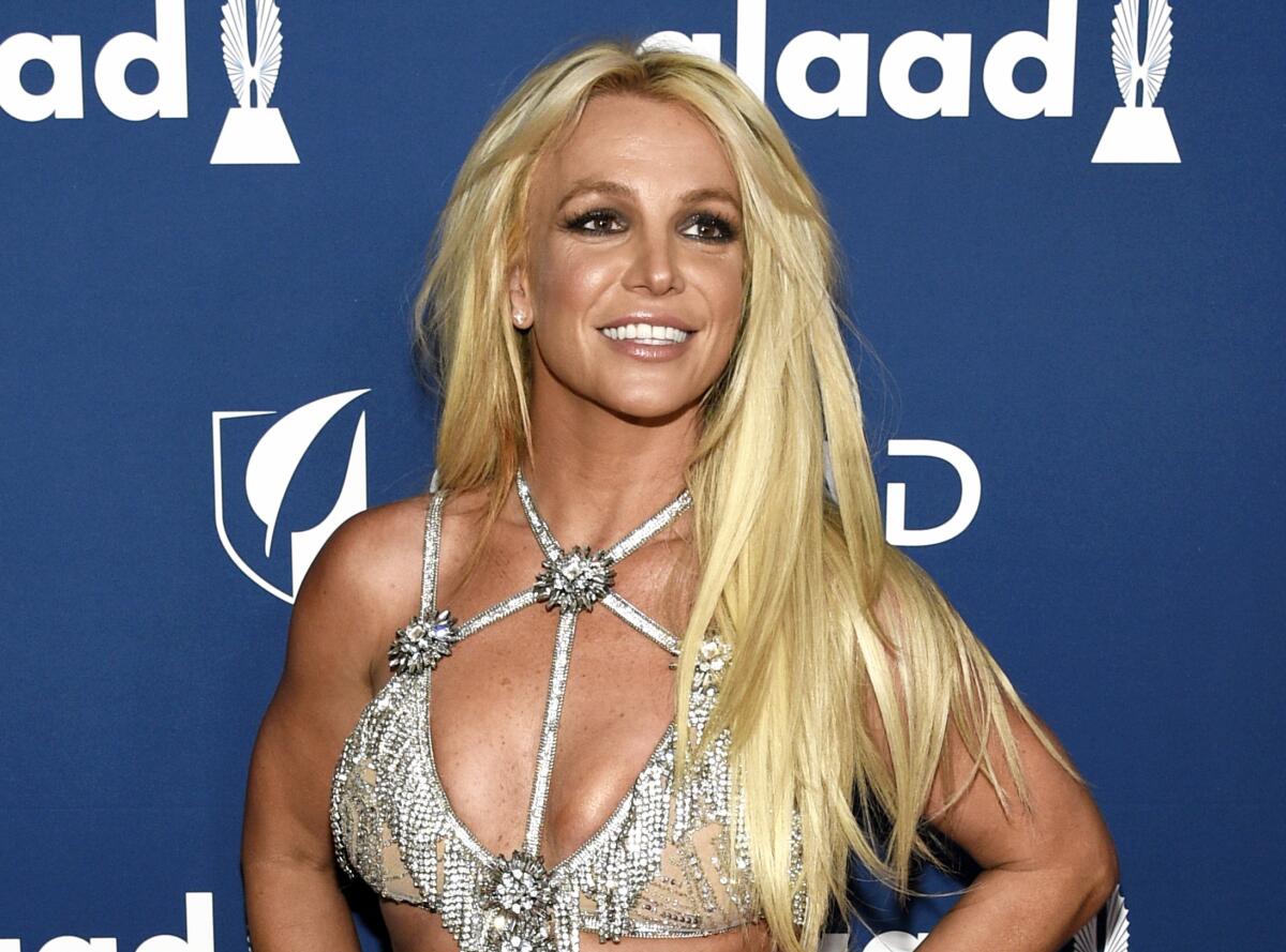 Britney Spears with long hair and a silver, strappy bra top smiles in front of a blue backdrop
