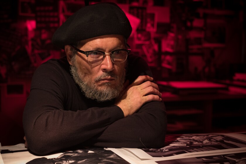 a man in a beret and glasses leans on a table with prints on it 