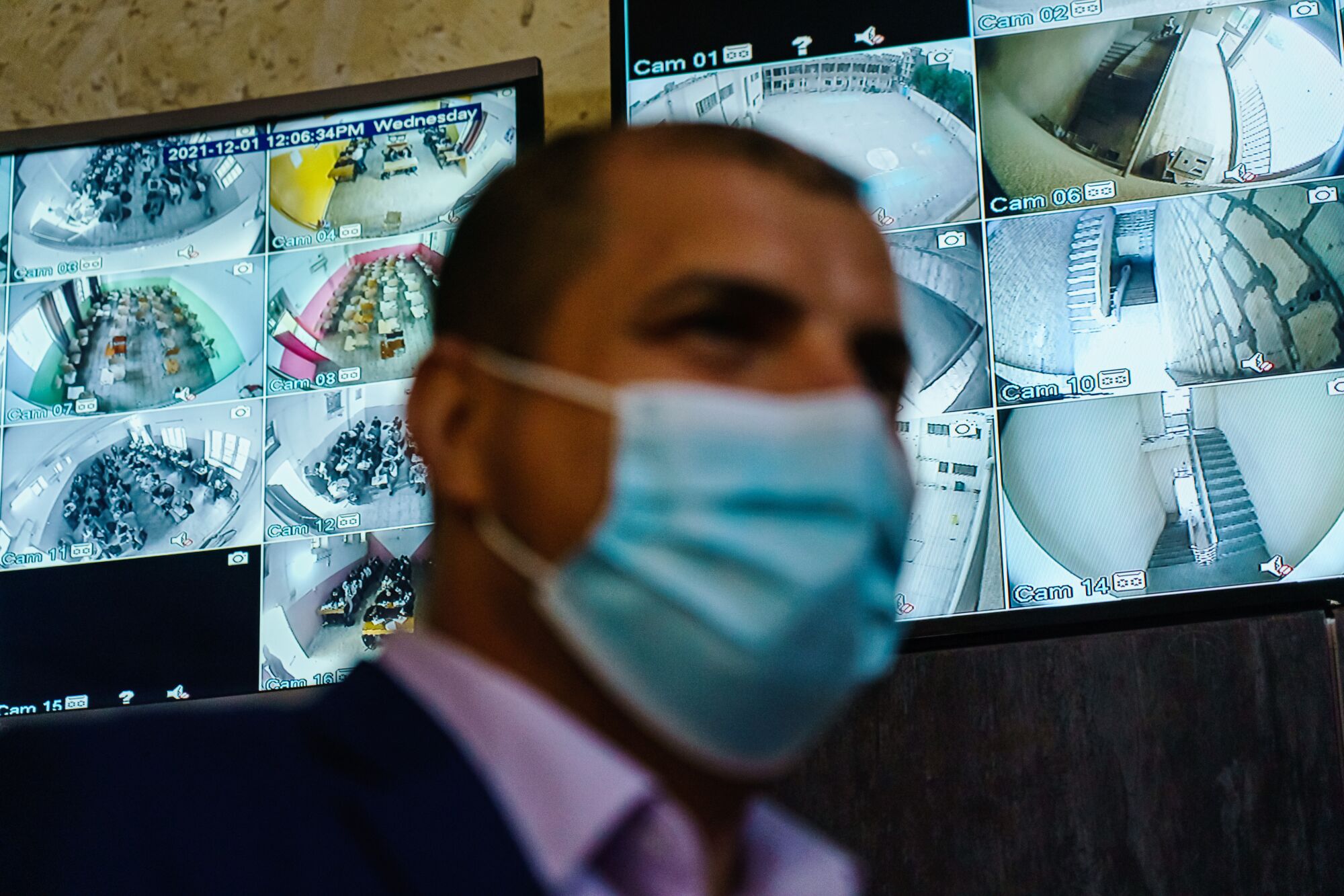  A man wearing a blue mask is shown with closed-circuit surveillance TV screens in the background