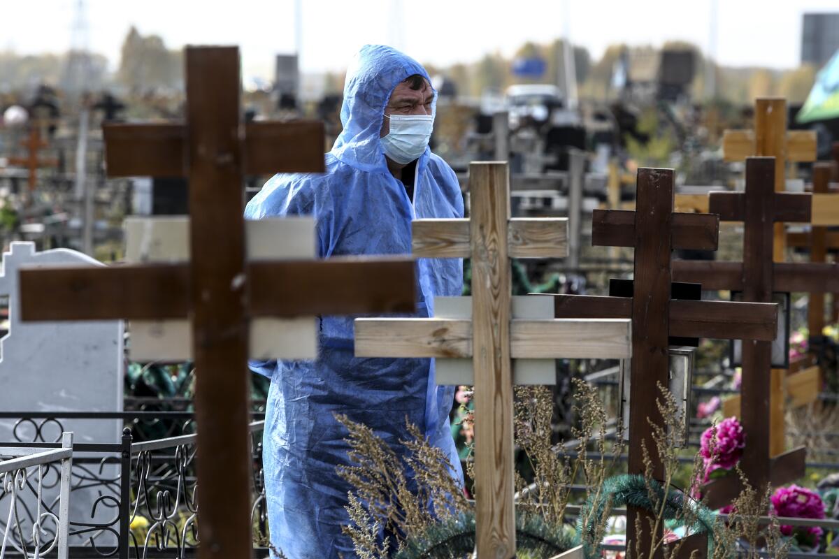 A masked man in a blue protective suit stand amid markers in a cemetery