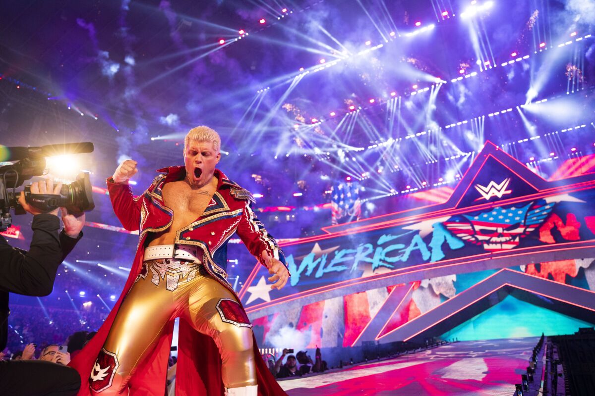 Cody Rhodes, dressed in a long red coat and gold pants, pumps his fist.