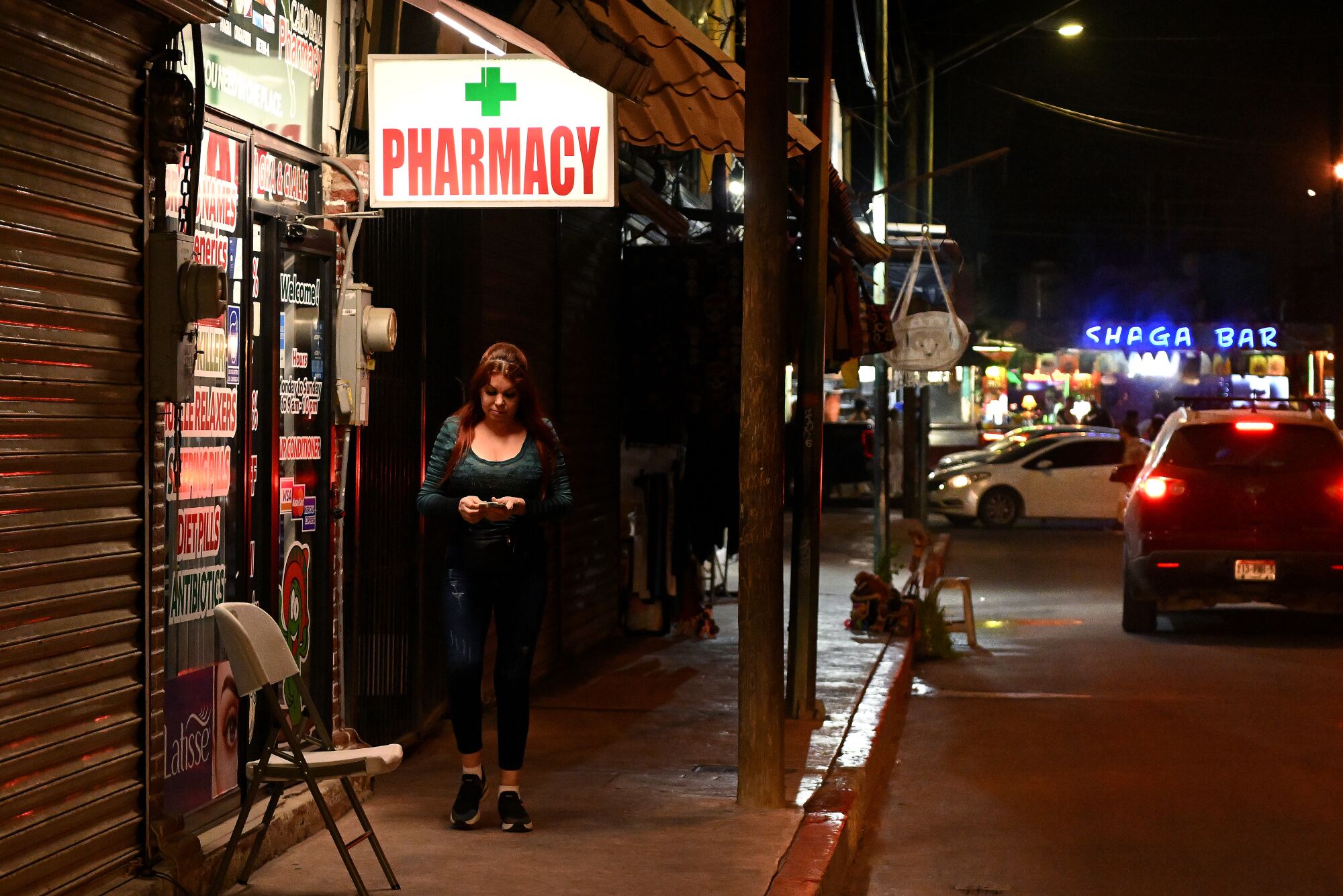 A woman passes by a store with a lighted "Pharmacy" sign at night while a car drives down the street next to her.