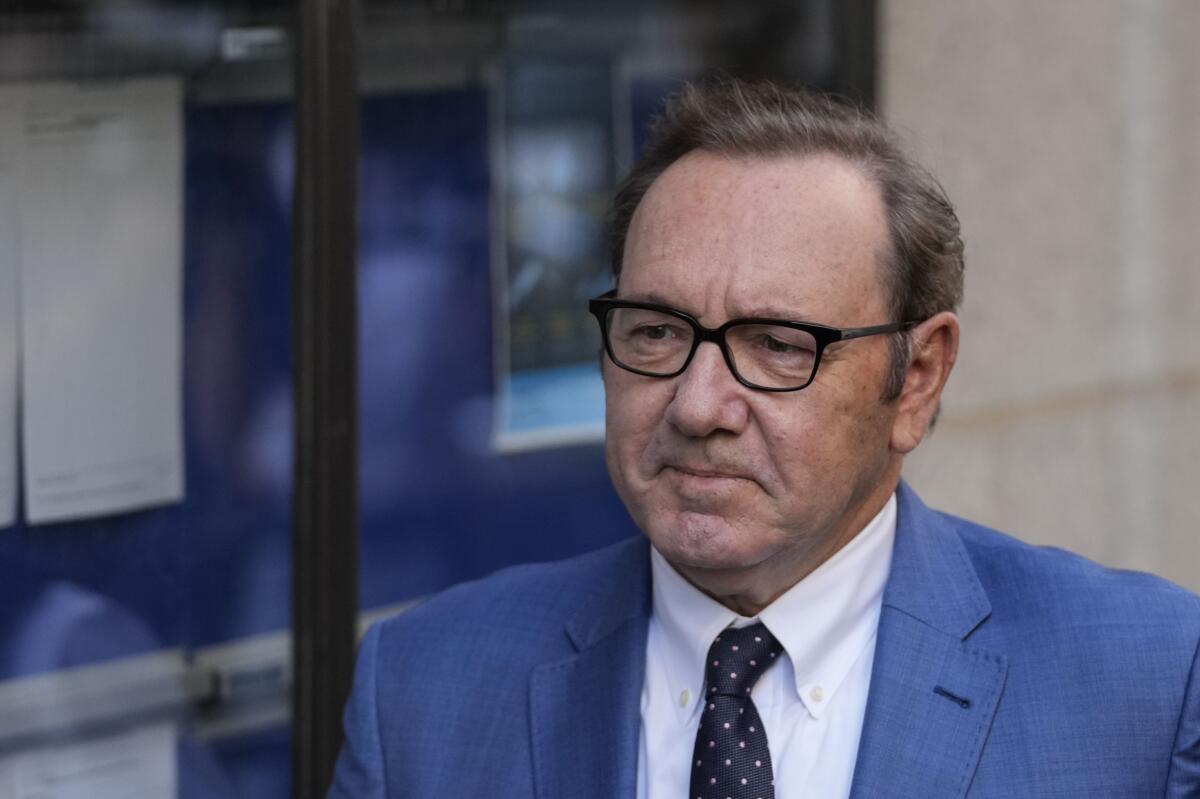 A man with short brown hair and glasses wearing a blue suit and polka-dot necktie