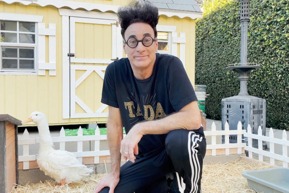 A man in sweatpants and round glasses kneels next to a duck in front of a shed 