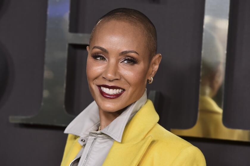 A woman with a shaved head smiling in dark red lipstick and a yellow jacket