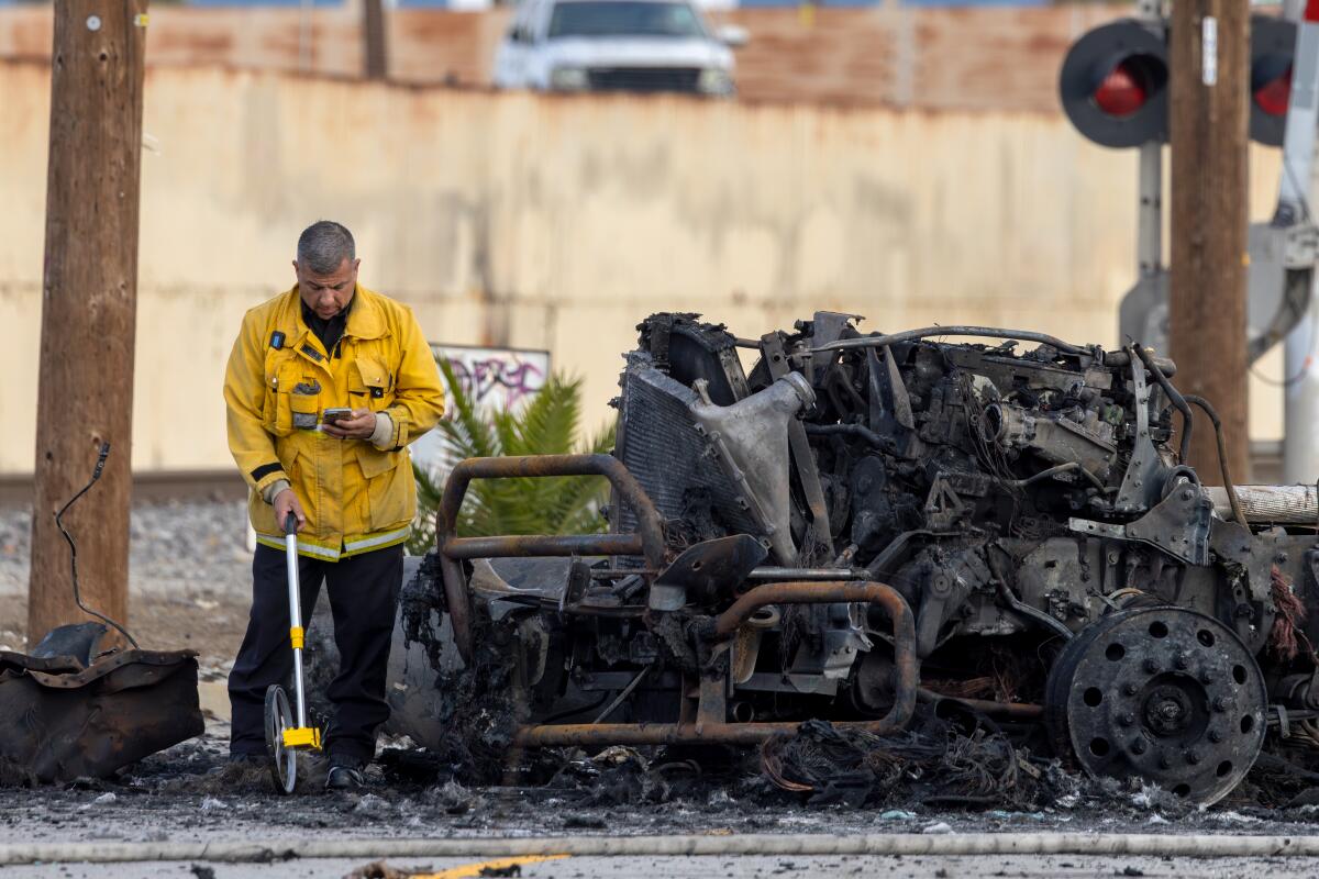 An investigator in a yellow reflective coat stands alongside mangled, burned wreckage of a truck.