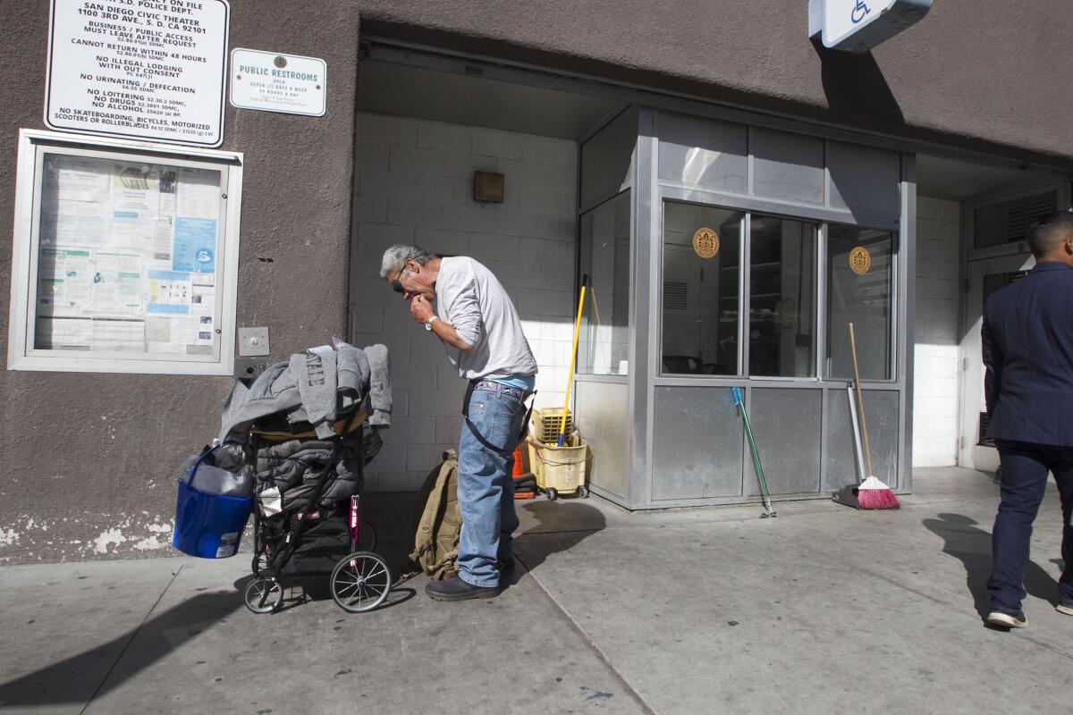 A man stands on a sidewalk near a building, stooped over a push cart piled with his belongings.