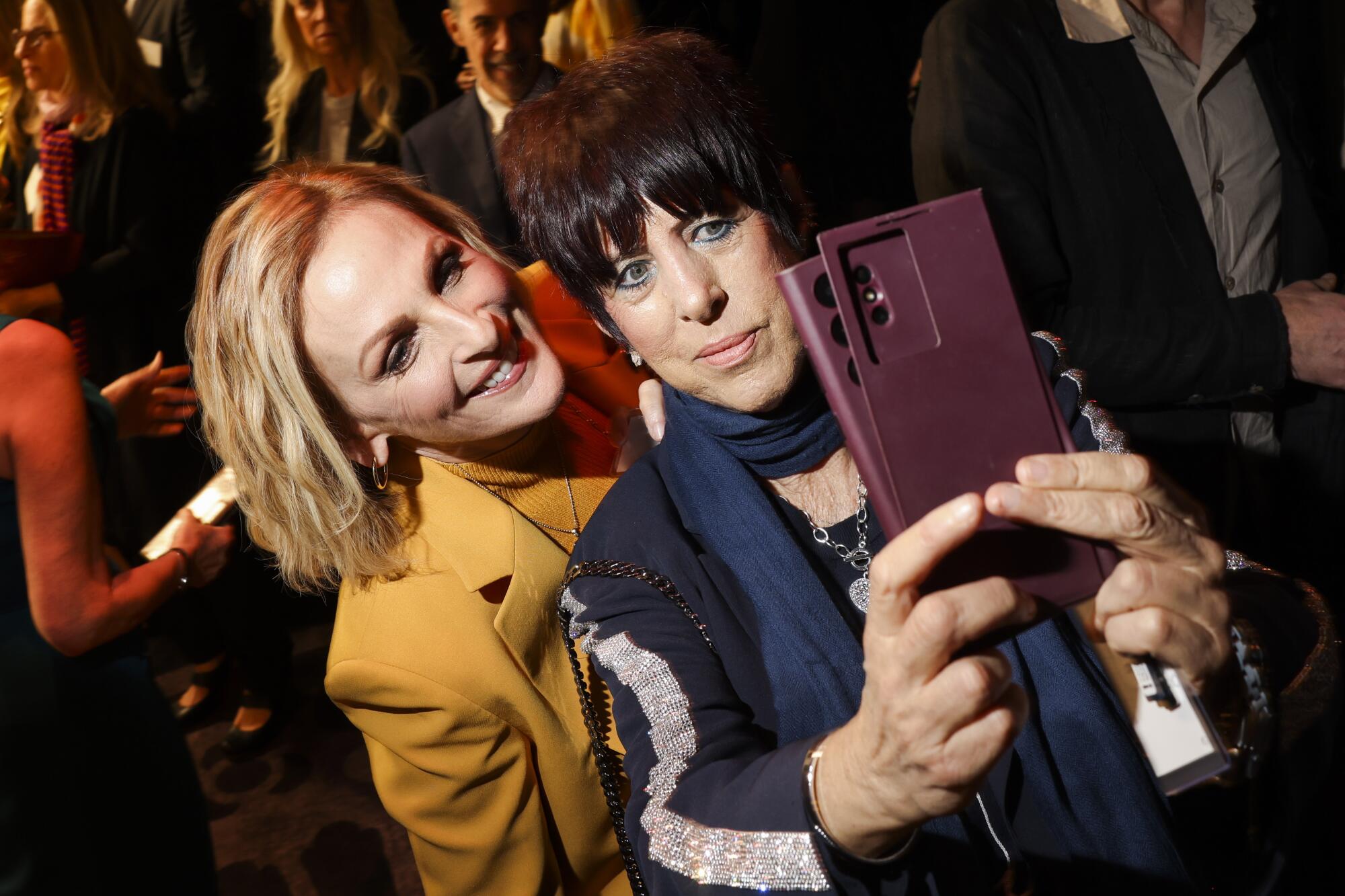 A blond woman stands behind a brunette woman as they pose for a selfie.