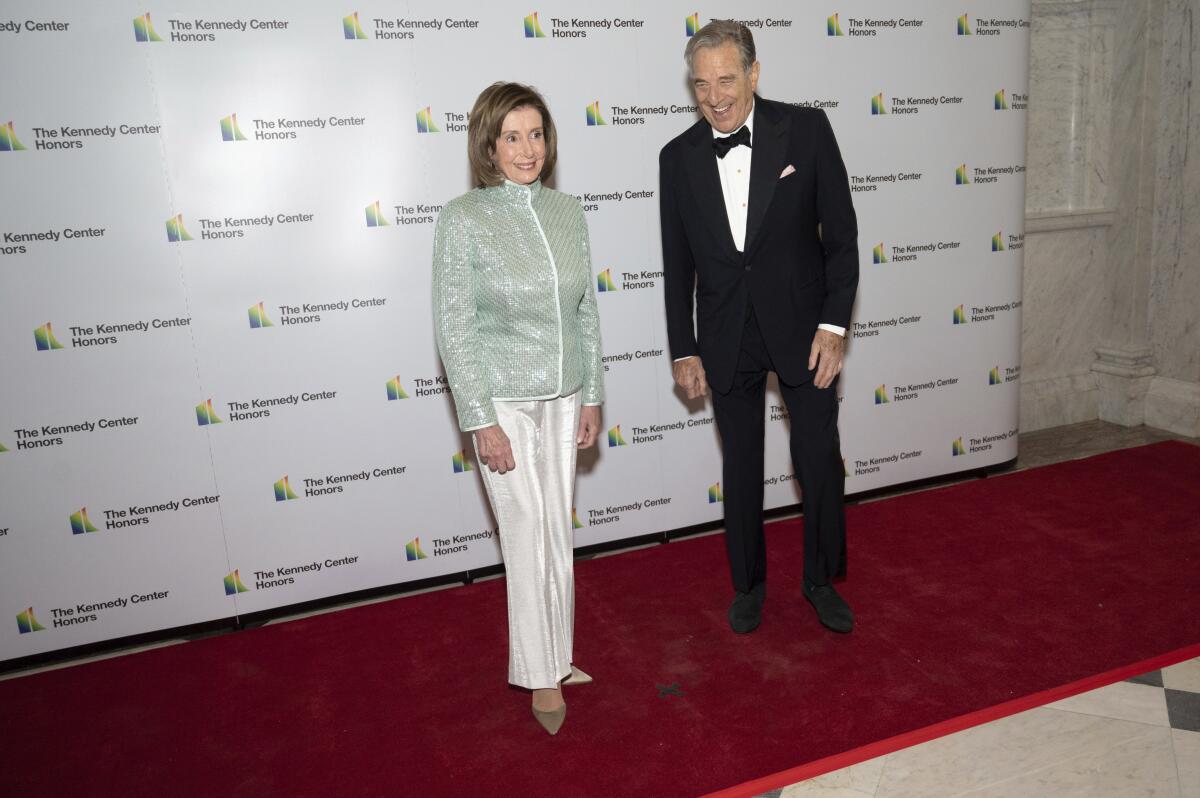 A man in a tux and a woman in a silky pantsuit stand on a red carpet.