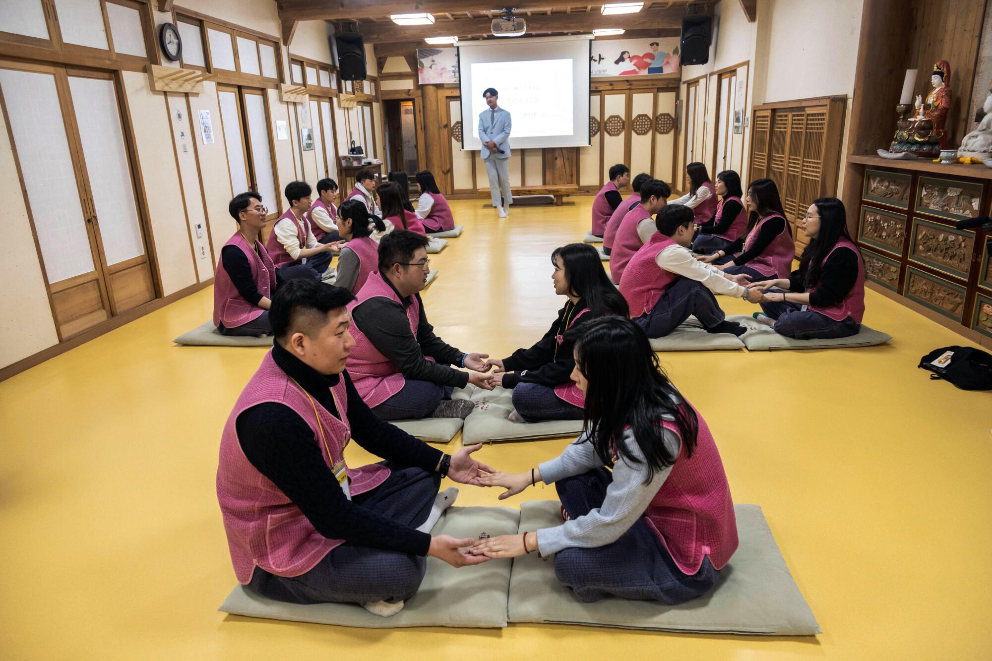Couples wearing deep pink vests, seated on mats facing each other, hold hands 