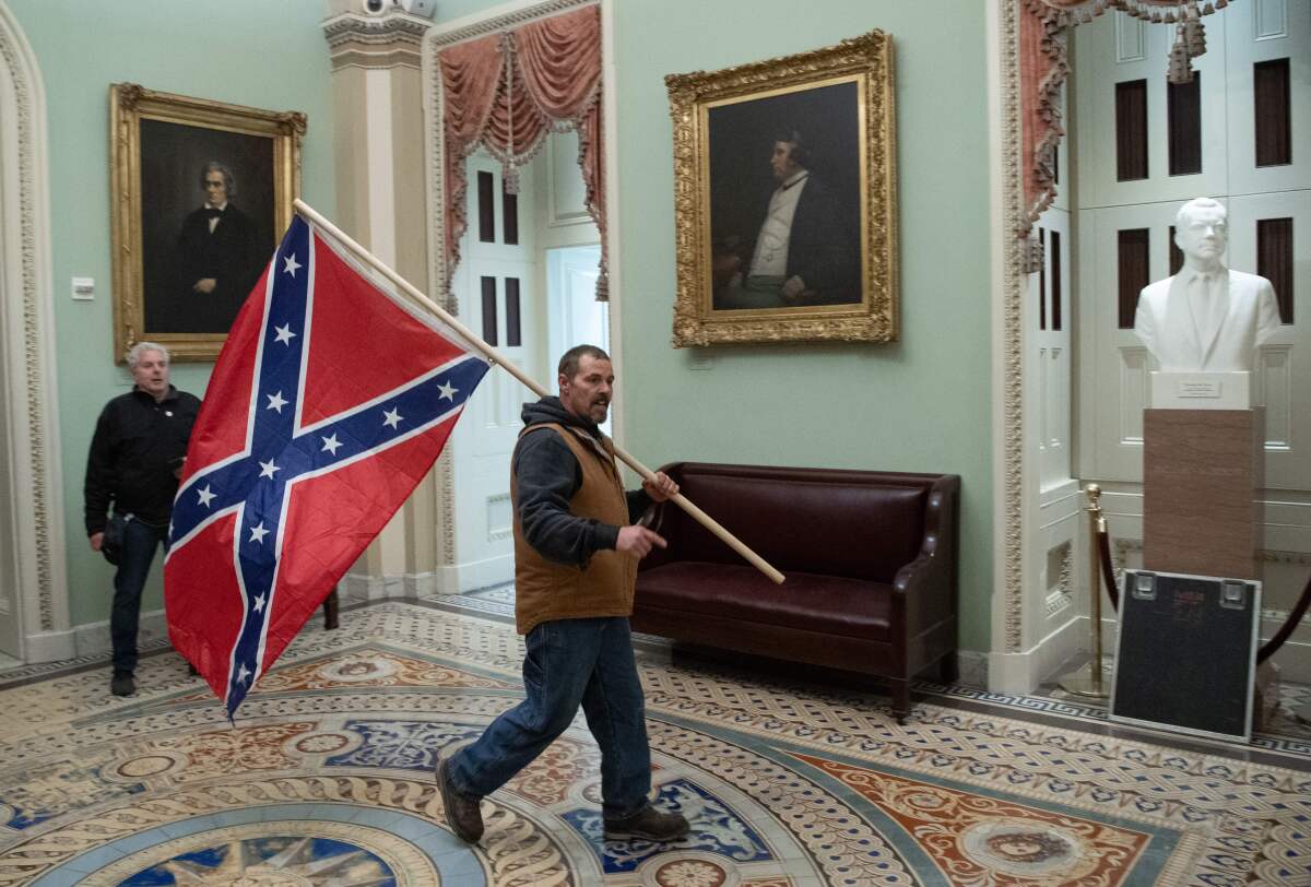 An extremist walks before a portrait of John Calhoun while carrying a large Confederate battle flag.