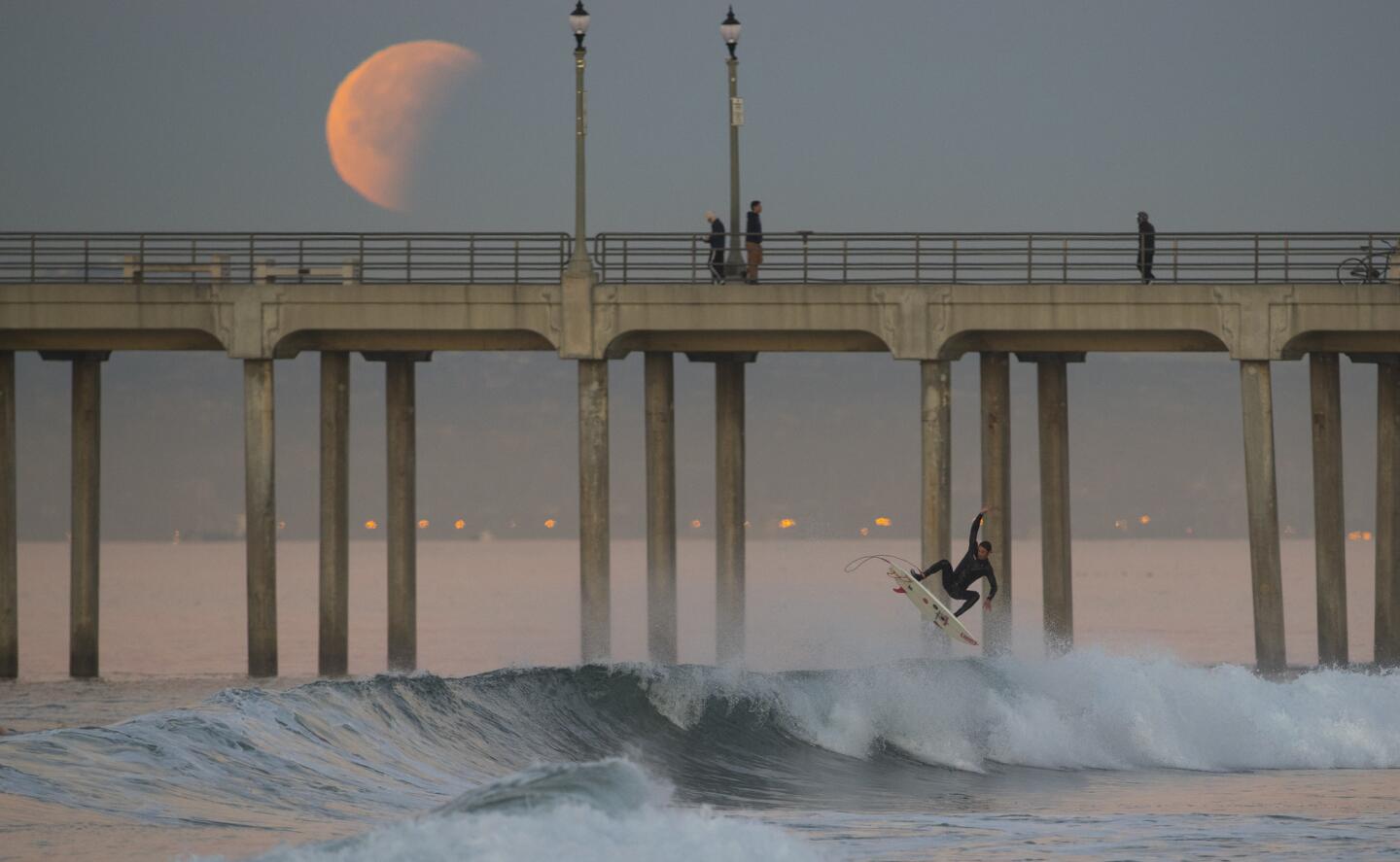 A surfer goes airborne off a wave as the super blue blood moon eclipse sets over the Huntington Beach pier.