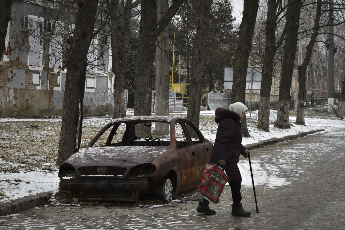 A person with a cane walks by a burned car on a snowy street.