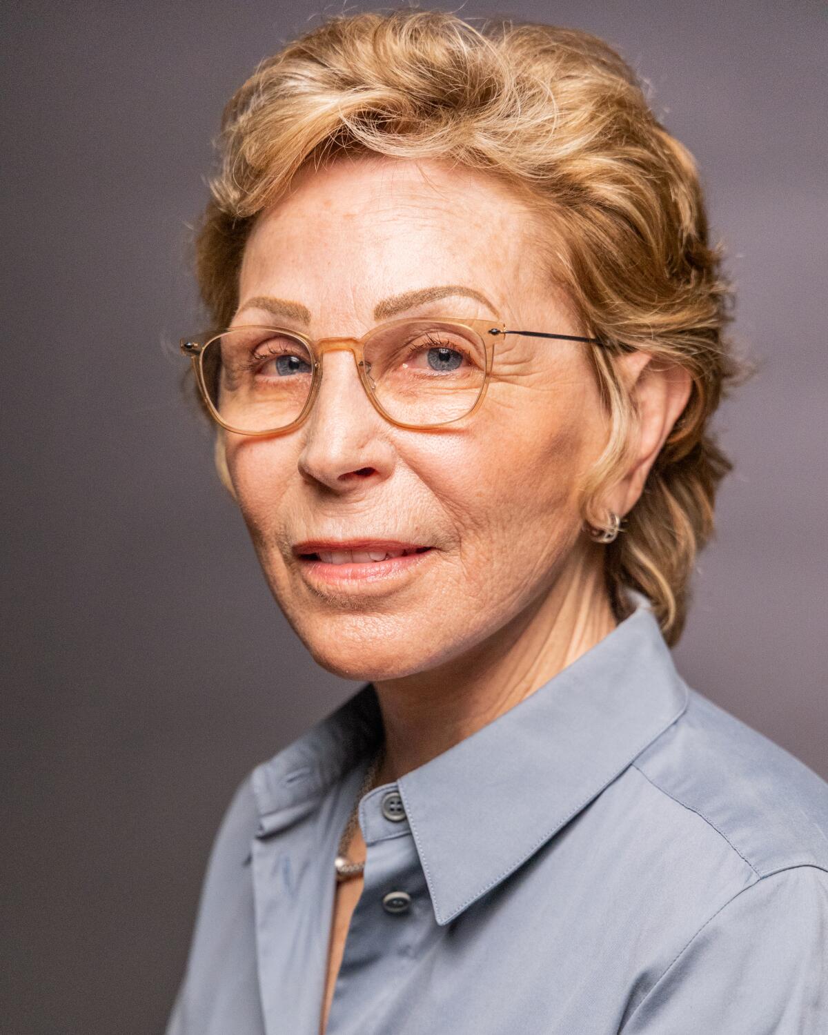 A portrait of a woman with short blonde hair and glasses wearing a gray collared shirt.