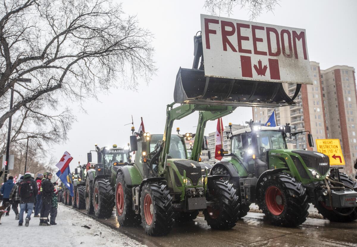Tractors are lined up on a city street. In the upraised scoop of one is a sign that says "Freedom" with a Canadian flag.