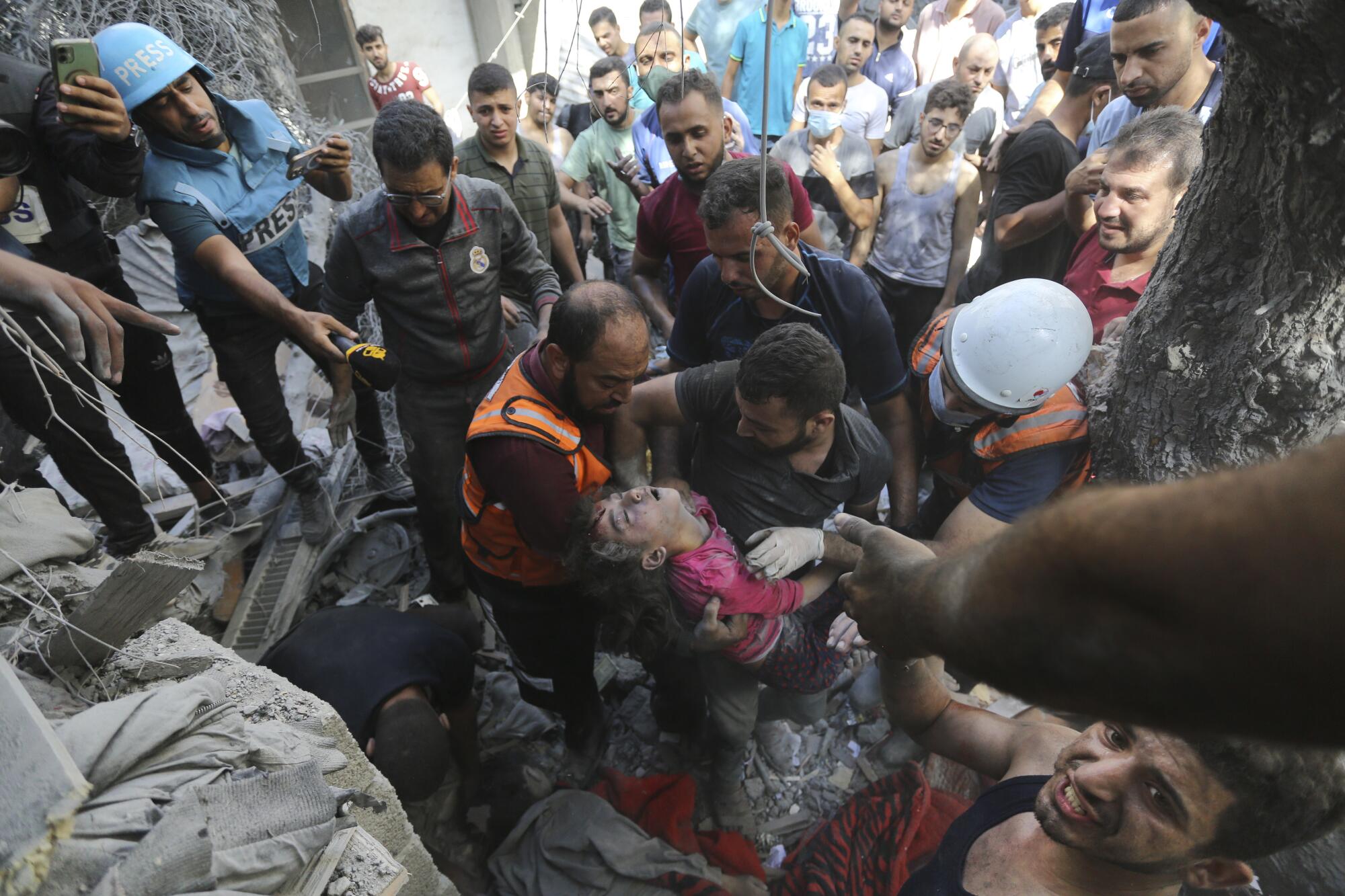 Two men hold a child in a red top amid rubble as a crowd looks on 
