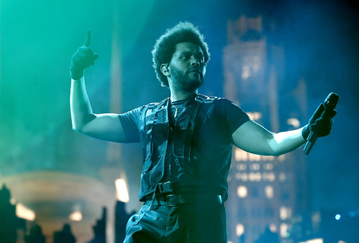 A man with an afro performs on a stadium stage wearing a tactical vest and black shirt, pants and gloves