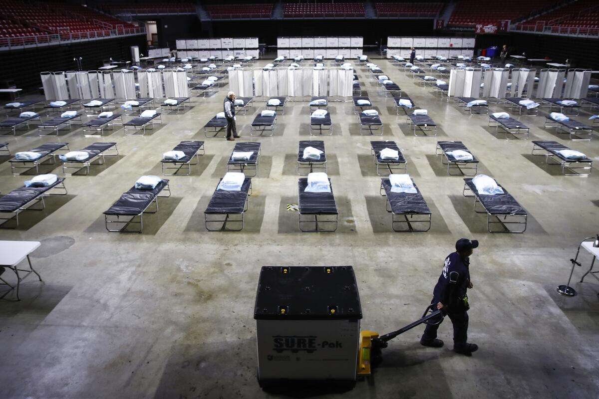 A worker pulls a pallet of supplies past rows of cots set up in a large, open building.