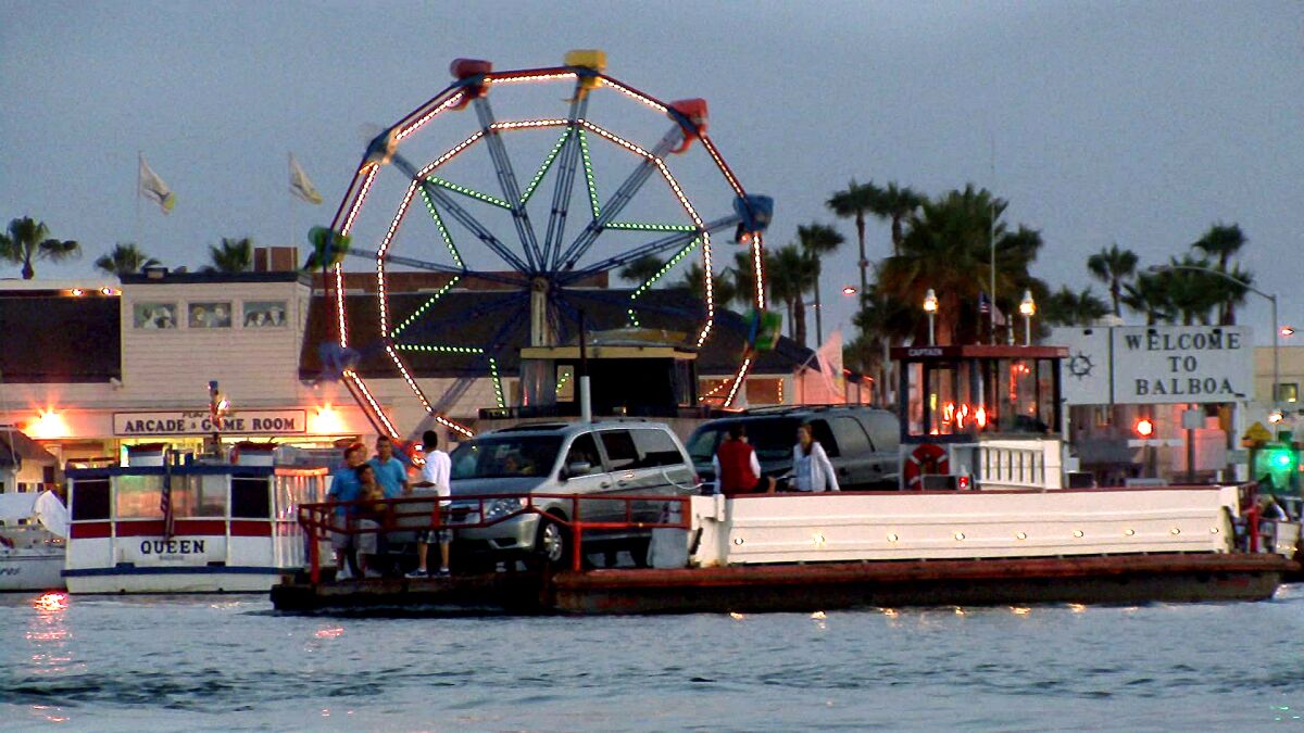 Cars and people on a ferry in front of a Ferris wheel and arcade building