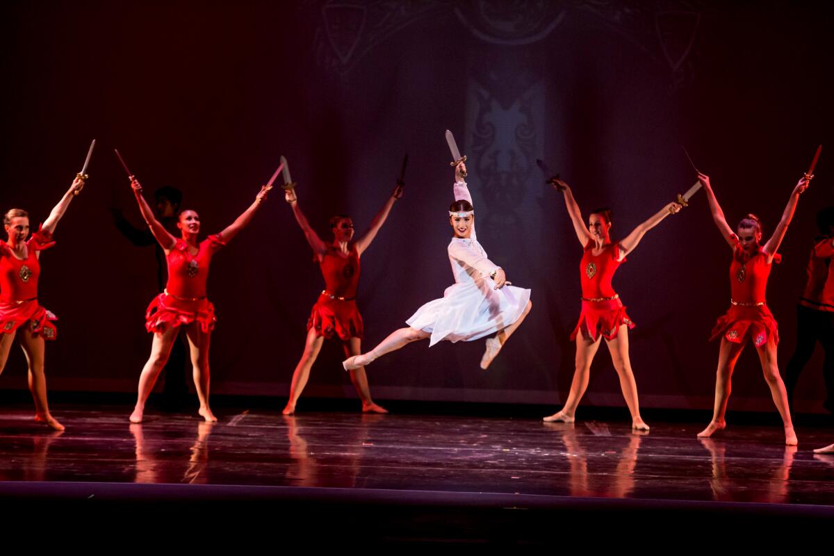 A dancer in white leaps into the air while others in red stand with arms and legs outstretched.