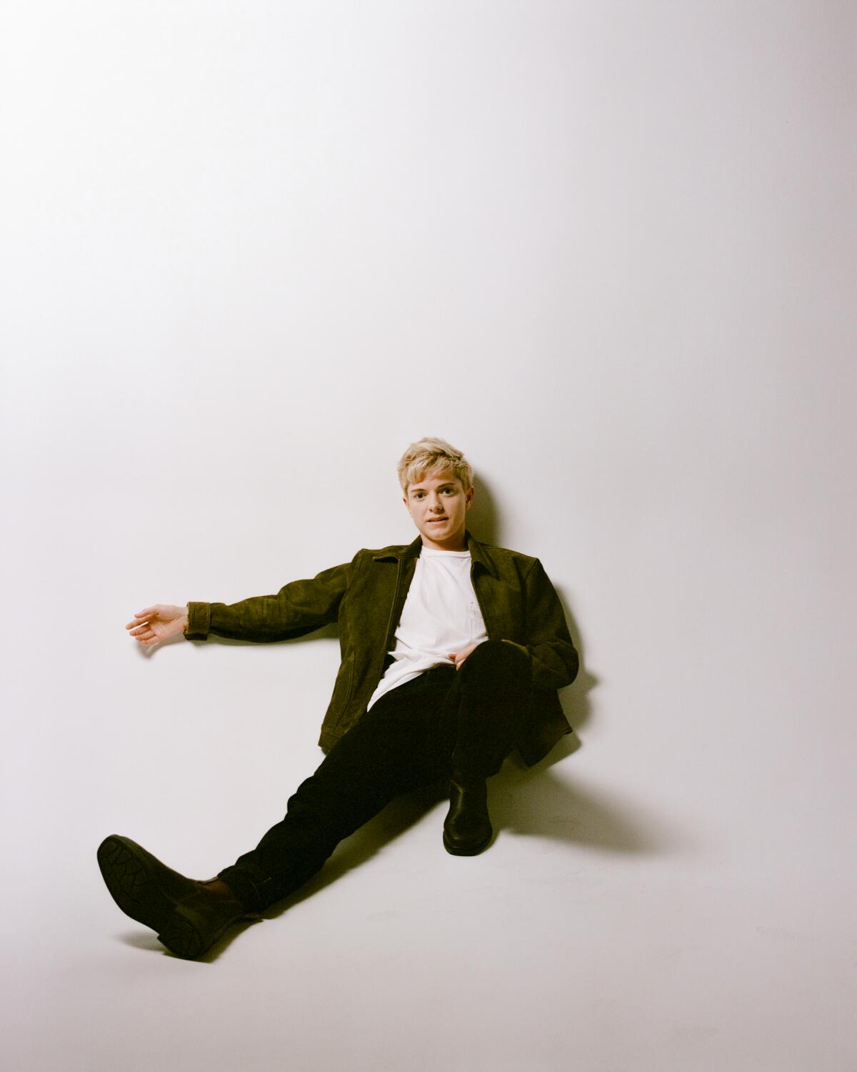 Comedian Mae Martin leaning against a wall with their legs outstretched. 