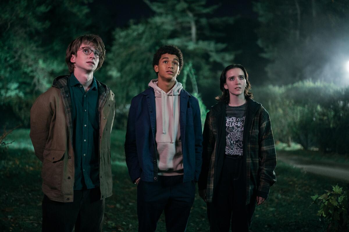 Two teenage boys and a girl stand in the dark in a wooded area. 