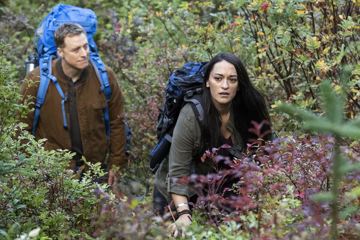 A man and a woman with blue backpacks walk through plants.