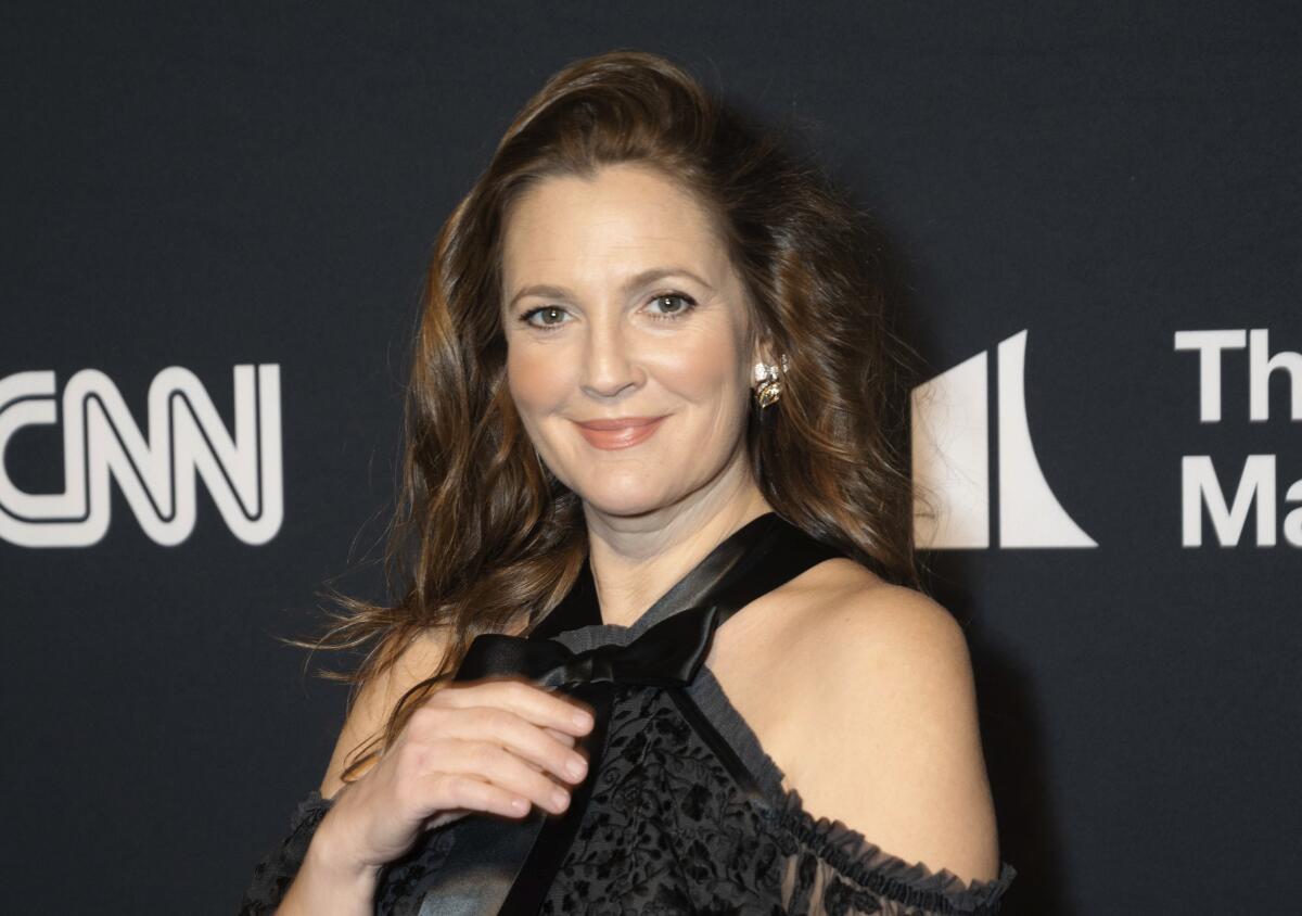 Drew Barrymore poses in a black, lacy, cold-shoulder dress in front of a black backdrop.