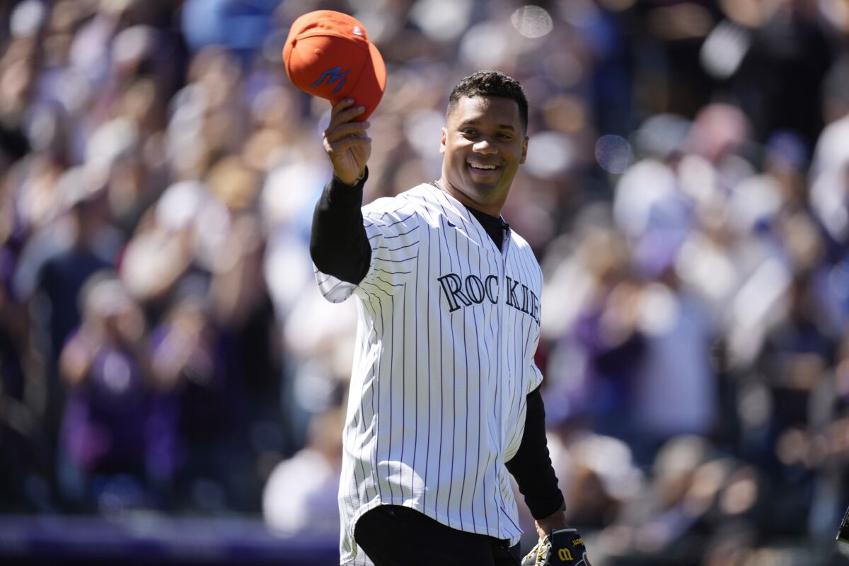 Denver Broncos quarterback Russell Wilson doffs his hat as he heads to the mound to throw out the first pitch during ceremonies to mark the regular-season home opener in a baseball game against the Los Angeles Dodgers Friday, April 8, 2022, in Denver. (AP Photo/David Zalubowski)