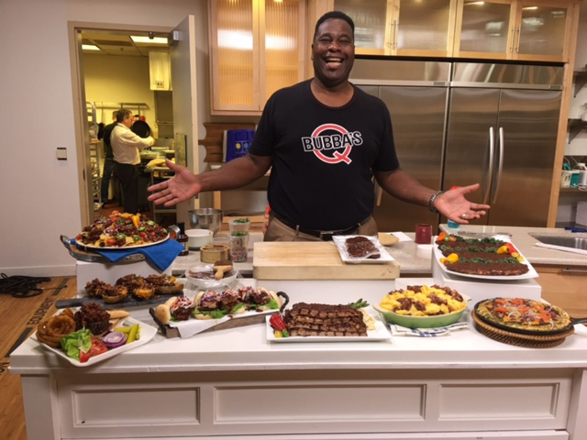 A smiling Al Baker gesturing toward a kitchen island filled with plates of food