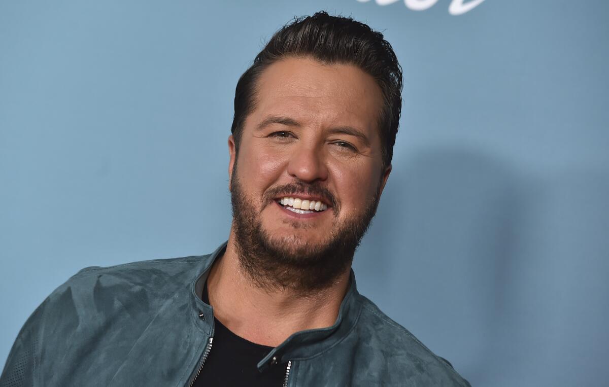 Luke Bryan smiles against a light blue background while wearing a light blue jacket