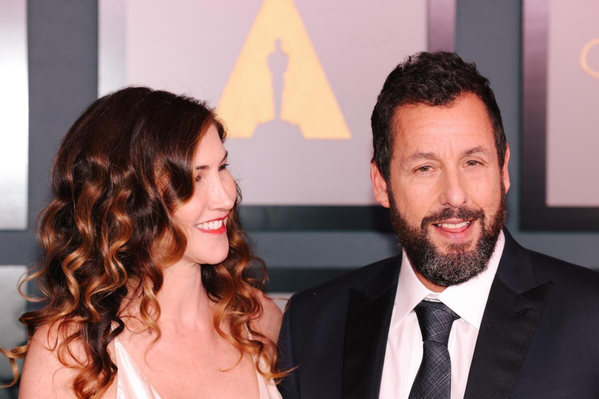 Jackie Sandler wearing a white dress smiles toward Adam Sandler who is wearing a black suit and tie